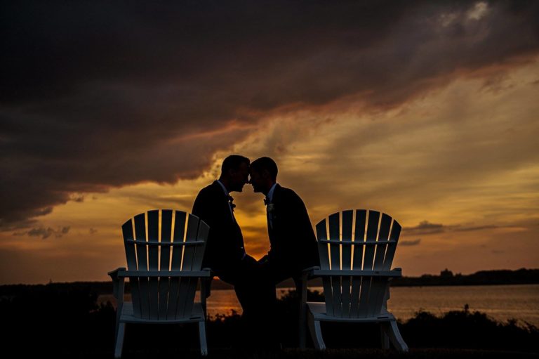 Two grooms kiss at sunset on Adirondack chairs at their wedding Castle Hill Inn Newport RI