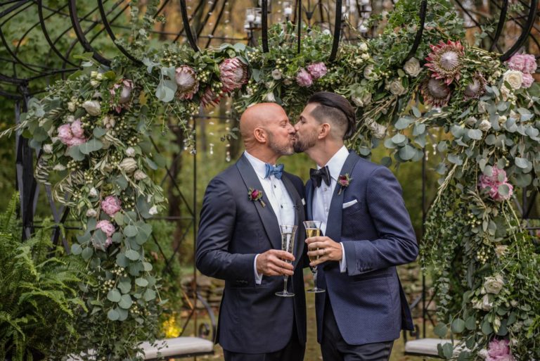 Two grooms kiss by floral arch Hana Floral at their wedding at Lord Thompson Manor CT