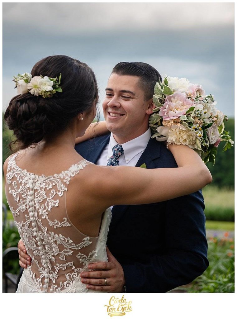 Smiling groom in J Crew suit hugs his bride in lace wedding dress at Tyrone Farm, Pomfret CT