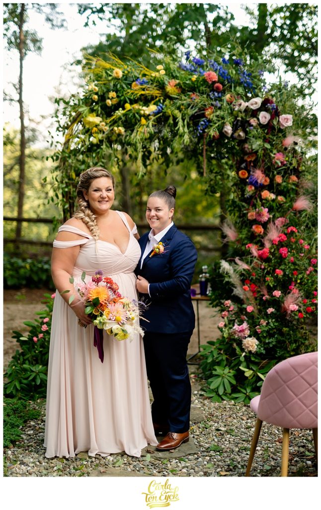 Two brides pose for photos near their rainbow ombre floral wedding arch in Weston CT
