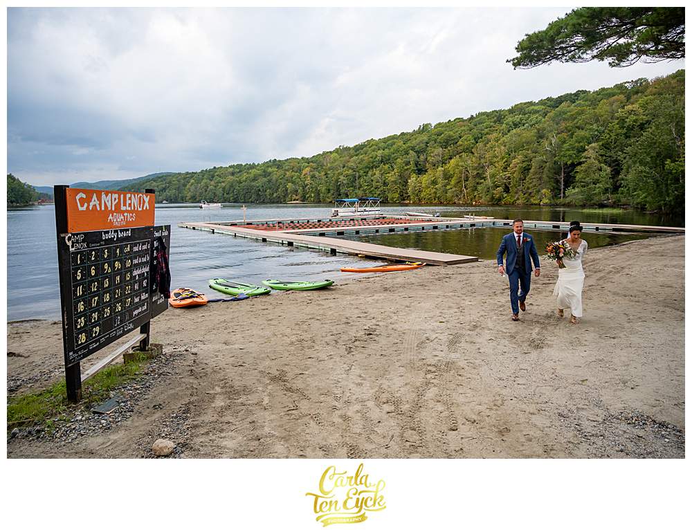 A bride and groom walk by the lake during their summer camp wedding at Camp Lenox in Otis MA