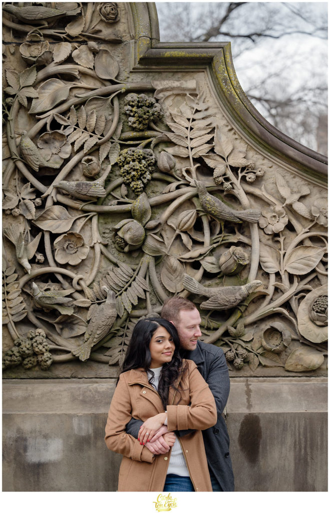 A couple cuddles by the stunning and iconic Bethesda Fountain in Central Park during their winter engagement session