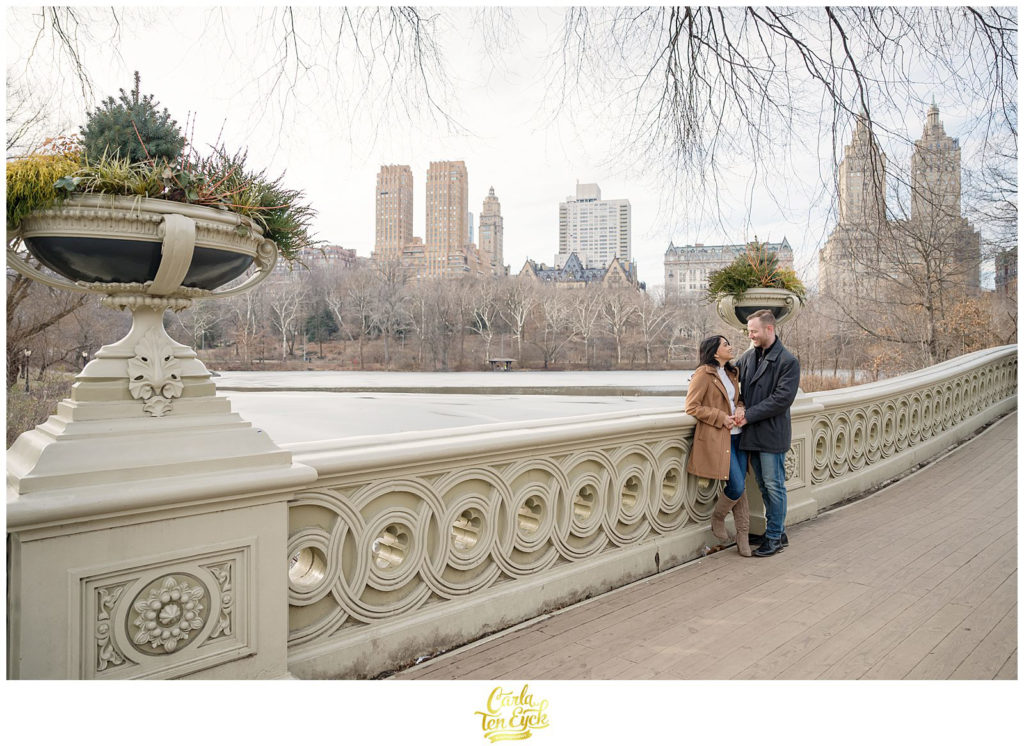 A couple laughs on the iconic Bow Bridge during their winter engagement session in Central Park NYC