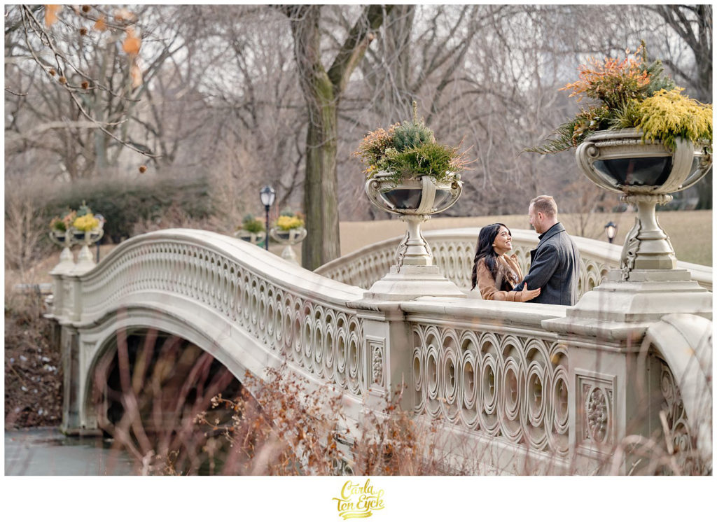 A couple laughs on the iconic Bow Bridge during their winter engagement session in Central Park NYC