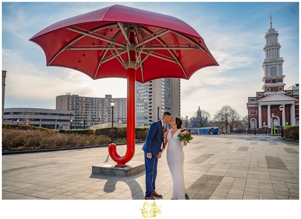 A bride and groom kiss underneath the red Traveler's umbrella in downtown Hartford at their Hartford City Hall Spring elopement, in Hartford CT